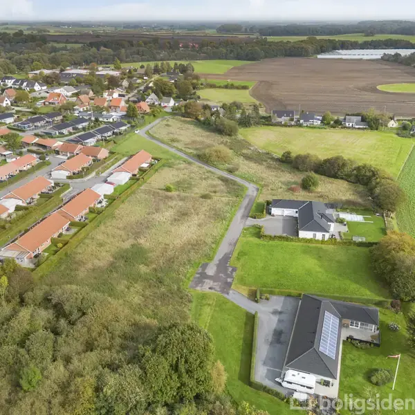 Boligkvarter med røde tage ses fra oven. Nogle huse har større grunde med grønne områder omkring. Bagved er marker og åbne landskaber.