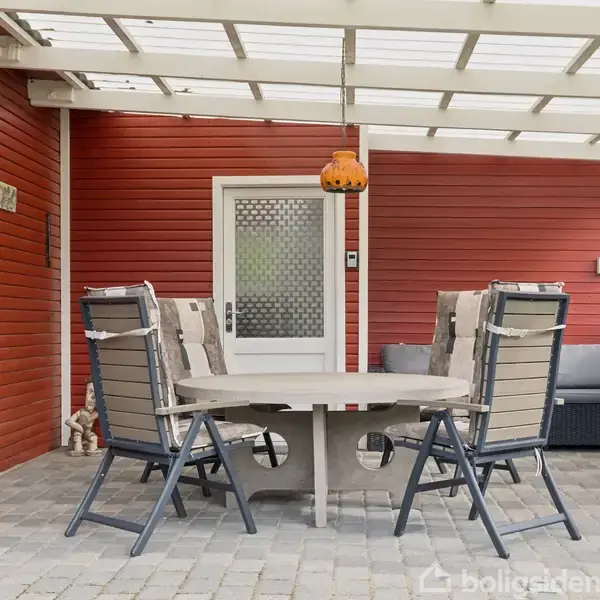 Terrassemøbler står rundt et rundt bord med tilhørende stole under en overdækket terrasse. Rød husvæg i baggrunden med en hvid dør og en farverig hængelampe tilføjer charme til scenen.
