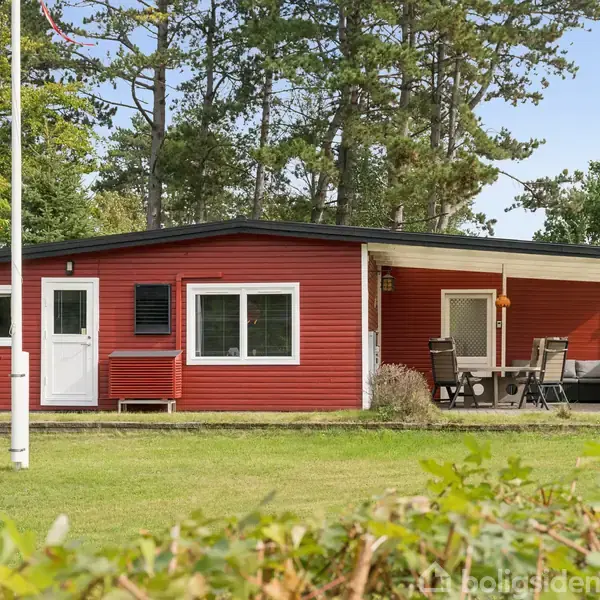 Rødt sommerhus står stille i skovklædt område. Rundt om er der græsplæne. Der er en overdækket terrasse med havemøbler til afslapning. En flagstang ses ved huset.