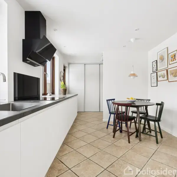 Et moderne køkken har en lang, hvid bordplade med vask og emhætte over. I baggrunden står et rundt spisebord med fire stole, omgivet af lyse vægge og vinduer.