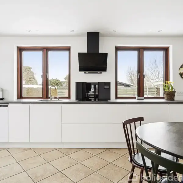 Et moderne køkken har hvide skabe, en sort emhætte, og to vinduer. På bordet står en skål med frugt. Lyset er klart, og gulvet har et skakternet mønster.