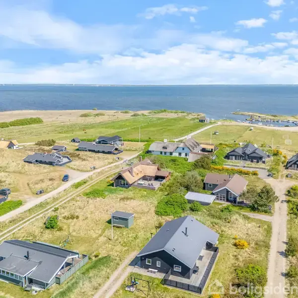 Flere huse ligger spredt i et landskab nær kysten, omgivet af græsmarker og veje, med havet i baggrunden under en skyfri himmel.