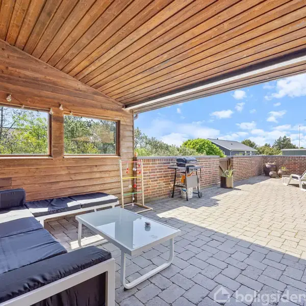 Overdækket terrasse med sorte sofaer og glasbord, hvor der er sollys gennem vinduerne. Udenfor er der solstole og en parasol på en brostensbelagt gårdhave.