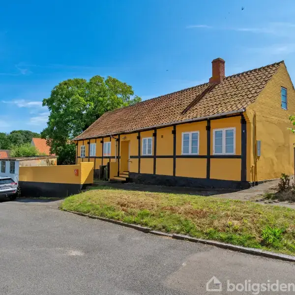 En gul bindingsværkshus med rødt tegltag står stille langs en vej. Flere biler er parkeret på asfalten foran huset. Grønt løv omkranser ejendommen i solskin.