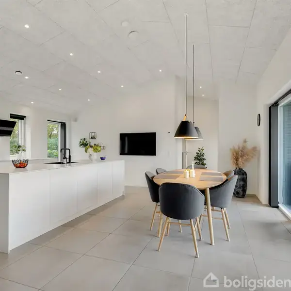 Køkken med hvid ø og sorte stole om et rundt spisebord. Hængende lamper over bordet. Store vinduer og moderne indretning. Grønne planter og lysindfald fra skydedøre i baggrunden.