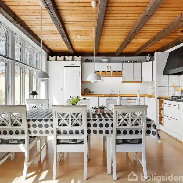 Et lyst køkken med trægulv og træloft. Et spisebord med polkaprikket dug og seks stole står centralt. Store vinduer til venstre og moderne apparater i baggrunden.