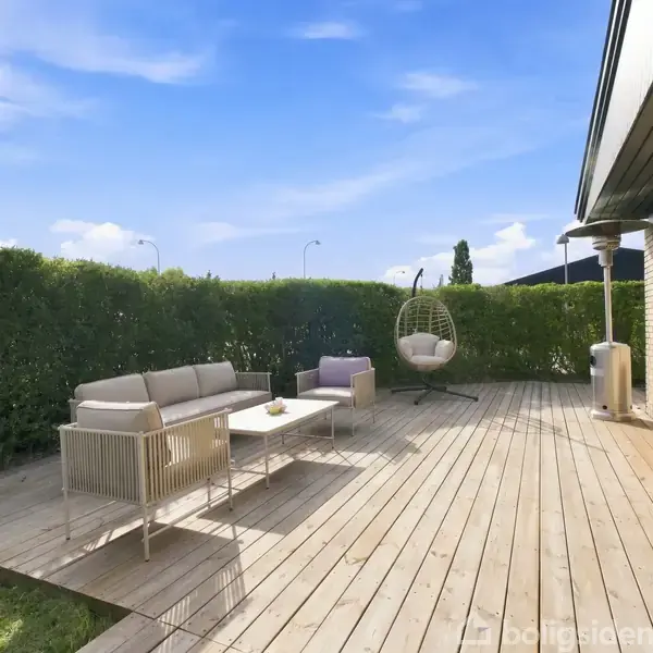 En terrasse i træ med en sofa- og hængestolssæt, placeres ved siden af en murstensbygning. Omkranset af grønne hække og en klar blå himmel ovenfor.