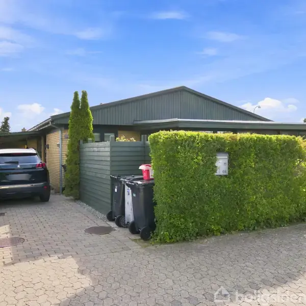 Et hus ligger bag en tætklippet hæk; en bil holder parkeret i indkørslen ved siden af skraldespande. Himmel med lette skyer danner baggrunden.