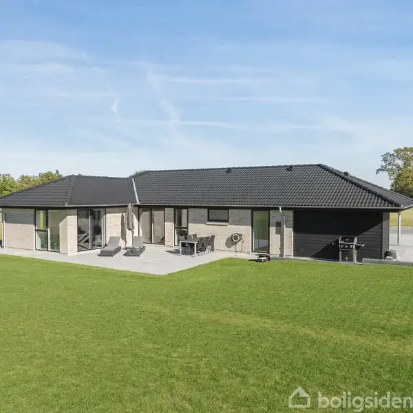 Én-etagers moderne hus med sort tag står på en grøn græsplæne, omgivet af åben natur. Der er en terrasse med havemøbler foran bygningen.