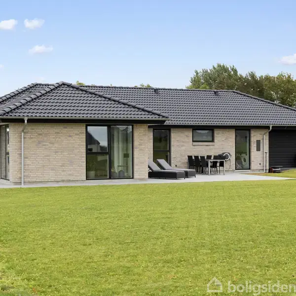 En moderne bungalow bygget af lyse mursten har store vinduer med sorte rammer og en terrasse med havemøbler. Den ligger i en velplejet, grøn have med træer i baggrunden.