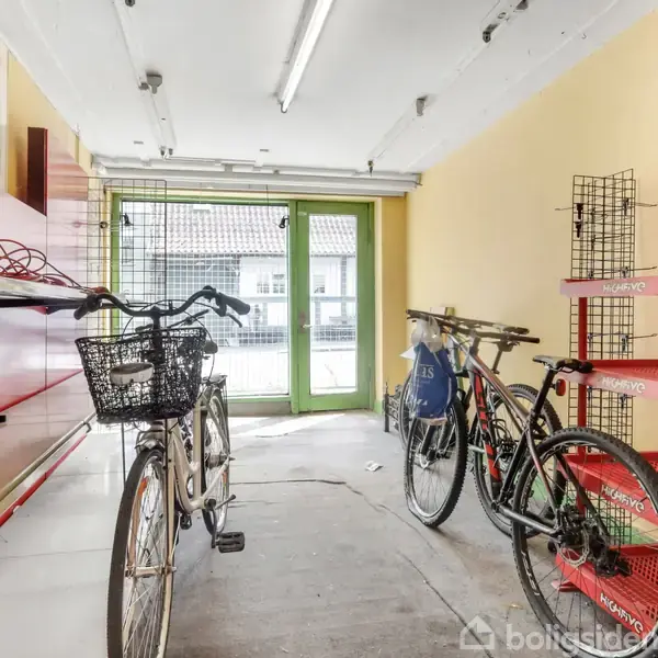 Cykler står parkeret i et indendørs rum med gule vægge. Til venstre er røde hylder, til højre en metalreol. Foran ses en stor grøn dør, der giver udsigt til huset udenfor.