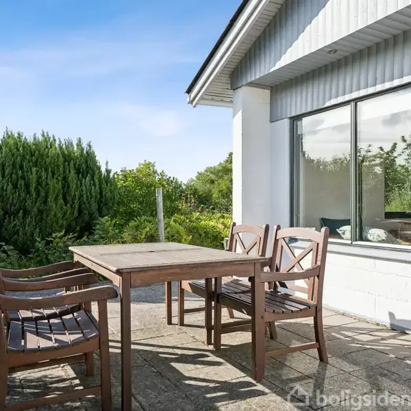 En terrasse med et træbord og fire stole, der står stille. Omgivet af en frodig have med høje buske. Flisebelagt gulv og udsigt over naturen gennem husets store vinduer.