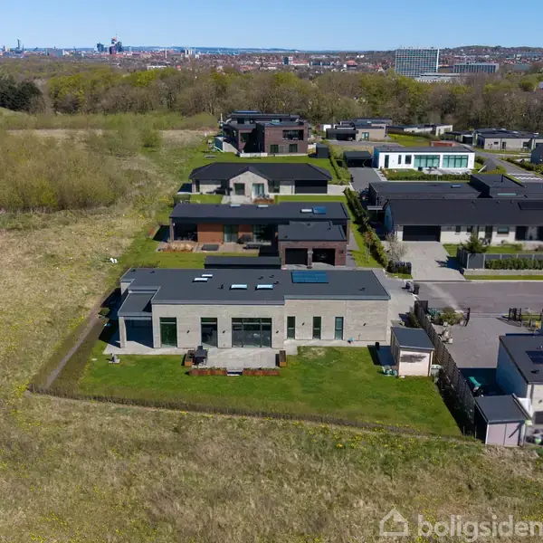Moderne huse står stille på række i et forstadsområde omgivet af græsmarker og træer, med en by i horisonten.