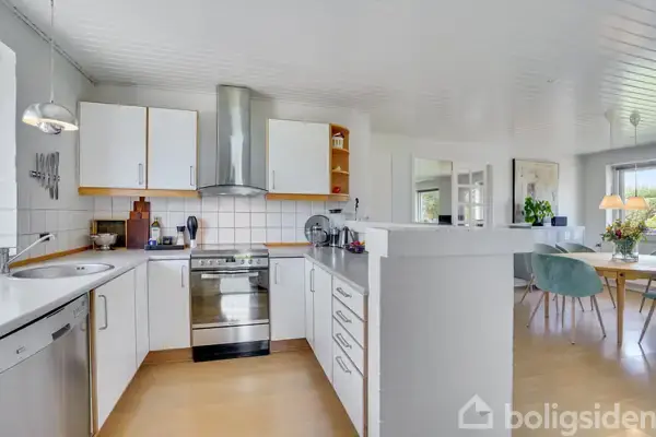 Et moderne køkken med hvide skabe og en stålkomfur, ledsaget af en emhætte. Køkkenet er lyst, og i baggrunden ses et spisebord med blomsterdekorationer.
