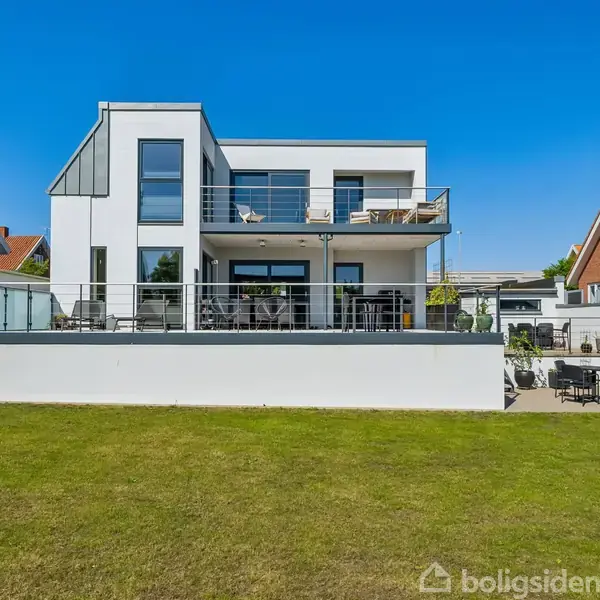 Moderne hvidt hus med store vinduer står stille på en grøn græsplæne. Huset har en rummelig terrasse og altan på første sal. Nabohuse kan ses på begge sider.