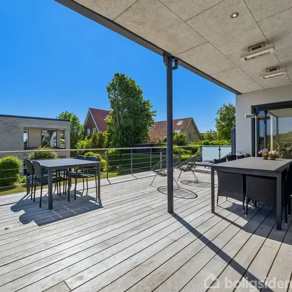 En udendørs terrasse med spisebord og stole, med udsigt over en velplejet have og flere huse i baggrunden. Skinnende blå himmel og sollys skaber en lys og åben atmosfære.