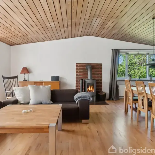 Stue med træmøbler og brændeovn, stående ved murstensvæg. Sofa og sofabord i forgrunden, spisebord med stole i baggrunden. Store vinduer lyser rummet op med udsigt til træer.