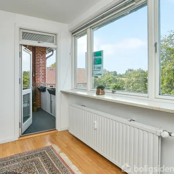Vindue åbner ud til en lille balkon med murstensvægge. Radiator står foran vinduet, hvorpå der er en lille plante. Udsigt til hustage og træer. Skilt: "Til salg".