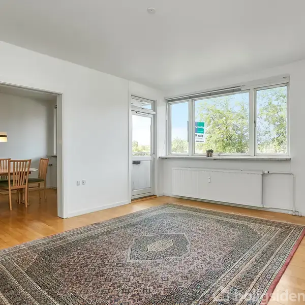 Et lyst rum med et stort tæppe på trægulv. En åbning fører til en spisestue med træmøbler. Store vinduer til venstre giver udsigt til træer.