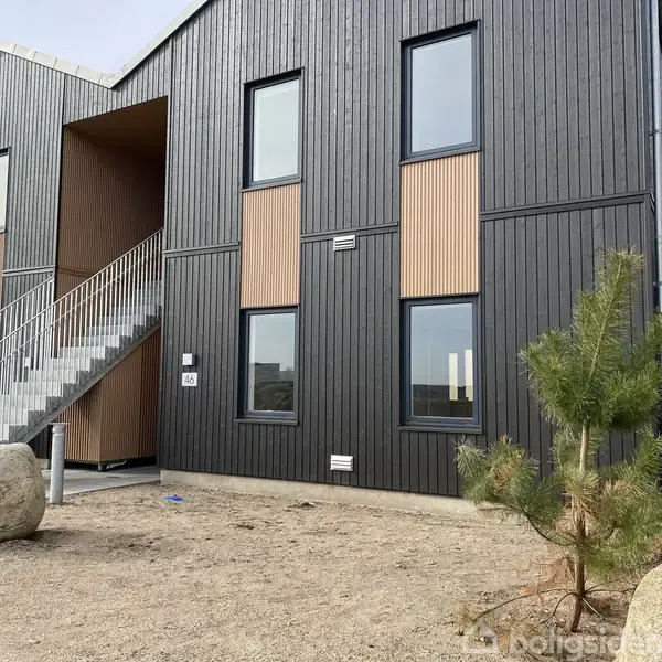 Bygning med mørke træfacader og store vinduer, der står på en grusbelagt grund med et lille træ og en sten. Trappe fører op til første etage. Husnummer 4G.
