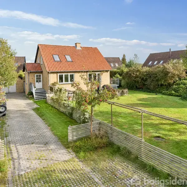 Huset er en murstensbygning med rødt tag, placeret i en have med græsplæne og flere træer. To motorcykler står parkeret på en flisebelagt indkørsel.