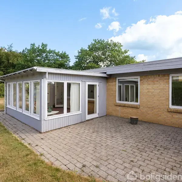 En murstensbygning med en glasudestue til højre står på en flisebelagt terrasse omgivet af en græsplæne og træer i baggrunden under en blå himmel.
