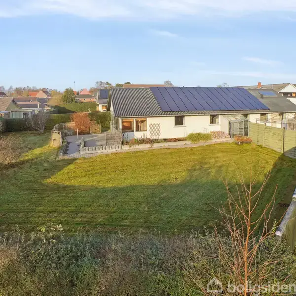 Et hus med solpaneler på taget står i centrum, omgivet af en stor græsplæne. Haven er indrammet af hegn og buske, med flere omkringliggende huse.