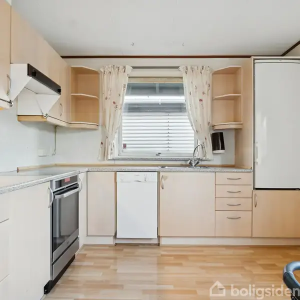 Køkken med lyse træskabe og -skuffer, komfur og emhætte til venstre, opvaskemaskine og vask under et vindue med gardiner i midten, køleskab til højre, lyse trægulve.