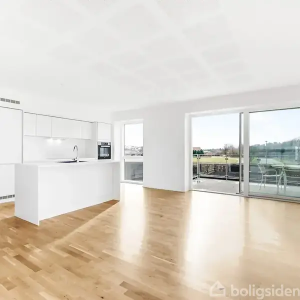 En lys og rummelig stue med åbent køkken. Køkkenet har moderne, hvide skabe og en indbygget ovn. Store skydedøre fører ud til en balkon med udsigt over et grønt område.