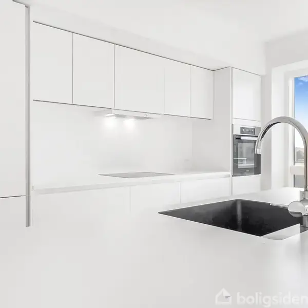 Et moderne, hvidt køkken med vask og vandhane. Lyset skinner ind fra et stort vindue med udsigt til en klar blå himmel. Skabe og ovn er minimalistiske og integrerede.