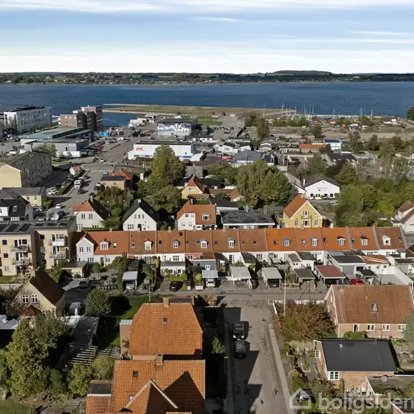 Bygningerne med røde tage ligger i en tæt række, omgivet af grønne træer. I baggrunden ses en havn ud mod en bred fjord, hvor flere både er fortøjet.