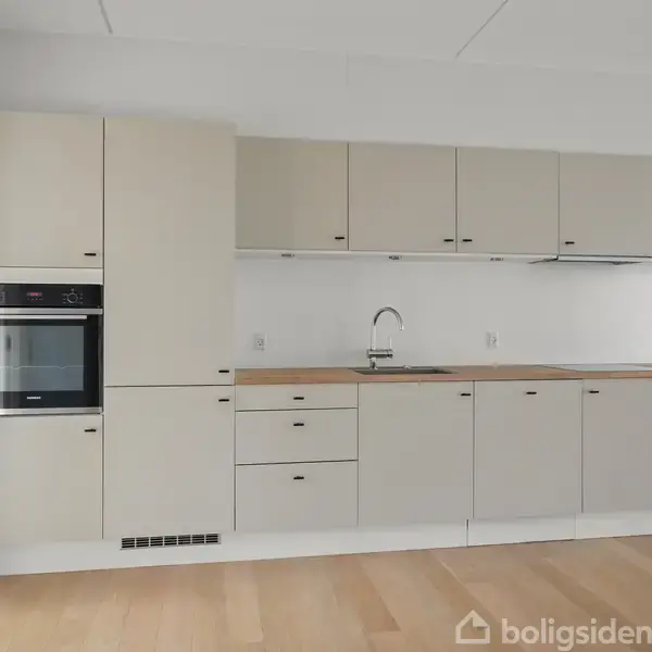 Et moderne køkken med beige skabe, en indbygget ovn, vask og emhætte. Det har lyse trægulve og hvide vægge. Rummet er minimalistisk indrettet med fokus på funktionalitet.