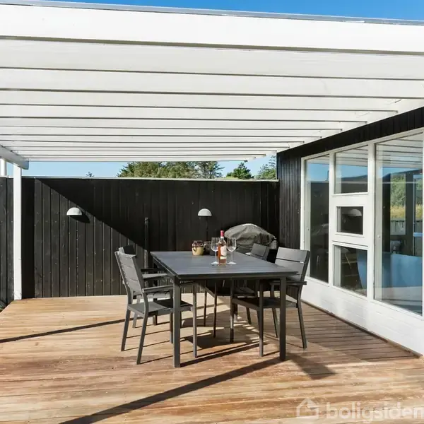 Terrassemøbler står på en træterrasse med en hvid pergola. Bordet har vin og glas. Mørke trævægge omkranser området, og glasdøre fører til et indendørs rum.