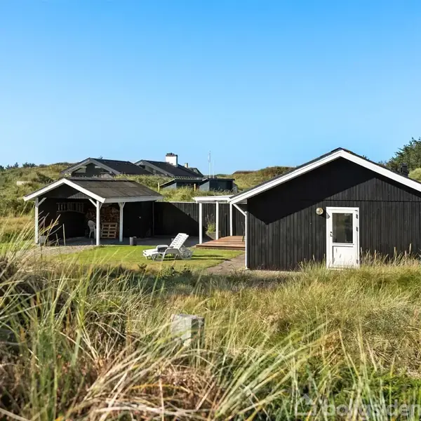 Et sort sommerhus står stille omgivet af højt græs på en åben mark. En terrasse med stole foran huset. Blåt, klart himmel fuldender den rolige scene.