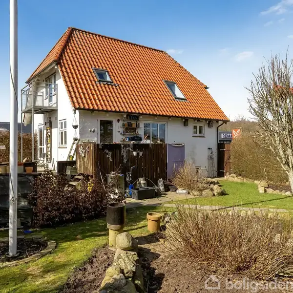 Et hvidt hus med rødt tag står i en have. Huset har flere vinduer og en flagstang i forgrunden. Haven er omgivet af buske og rummer buske og træer.