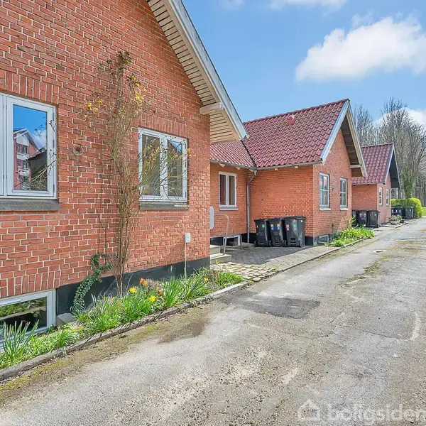 Rødt murstenshus med skråtag, placeret langs en smal vej. Huset har store vinduer, og der er blomsterbede foran. Andre huse og en hæk kan ses i baggrunden på en solrig dag.
