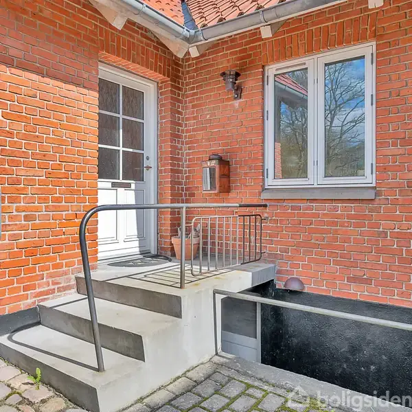 Murstenshus med hvid dør, to trappetrin og gelænder. Ved siden af er et vindue og en postkasse med teksten "HVL49". Omkringliggende belægning af stenfliser.