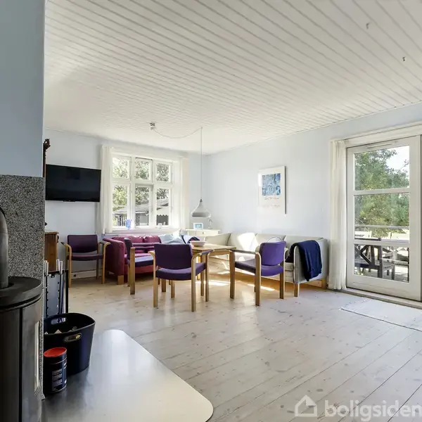 Stue med sofa- og spiseområde, hvor en brændeovn står ved siden af en køkkenbordplade. Store vinduer og en dør fører ud til en terrasse med udsigt til grønne omgivelser.