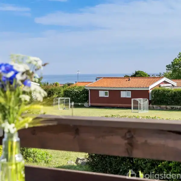 En vase med blomster står på en altan med udsigt til en rød bygning med rødt tag og små mål på en græsplæne. Havet er synligt i baggrunden.
