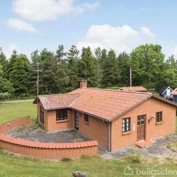 Rødt murstenshus med tegltag står omgivet af en græsplæne og omgivet af skov. Huset har en åben terrasse foran, omgivet af en halvmåneformet mur.