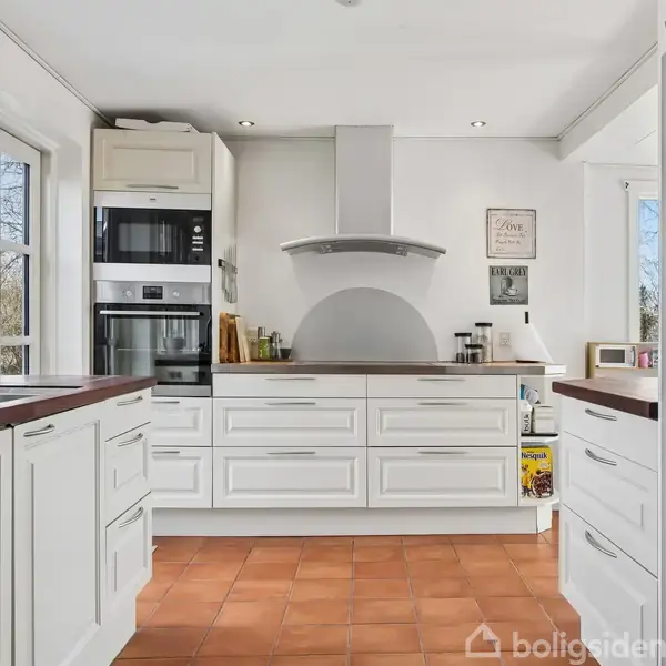 Et lyst køkken med hvide skabe og terrakottafarvede fliser. I midten ses en kogeø med indbygget komfur. På væggen hænger et skilt med teksten: "Love Kitchen Laugh Often."