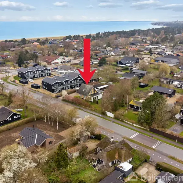 Rødt pil markerer et hus i et beboelseskvarter nær kysten. Huset, omgivet af andre boliger, ligger ved en vej med frodige træer i nærheden.