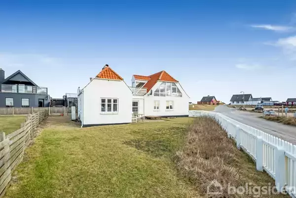 Et hvidt hus med rødt tegltag står i en græsplæne omkranset af et hegn. I baggrunden ses en vej og flere huse, alle under en klar blå himmel.