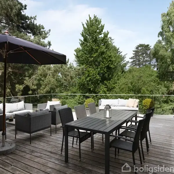 Terrassemøbler står på en bred træterrasse. En stor parasol skygger over et sofaarrangement. En spisebordsgruppe er placeret mod glasrækværket. Omgivet af frodig skovbevoksning.
