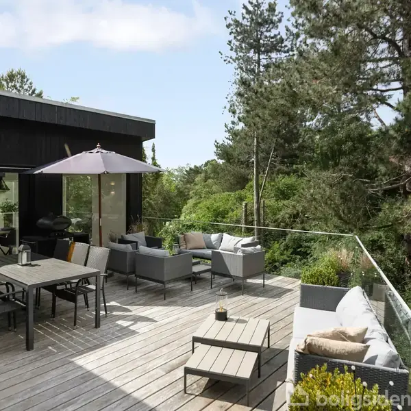 En træterrasse med spisebord og sofaarrangement under en parasol, omgivet af frodige grønne træer og buske, skaber en hyggelig udendørs atmosfære ved siden af et moderne hus.