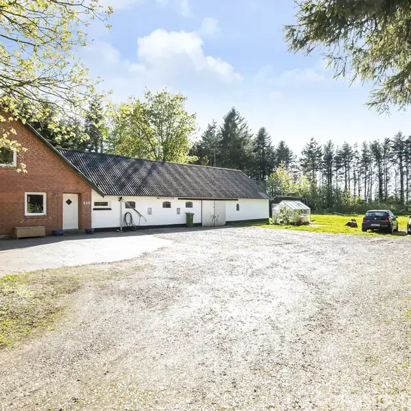 Et rødt murstenshus med sort tag står stille på en gårdsplads omgivet af træer og en skov i baggrunden. To biler holder parkeret til højre.