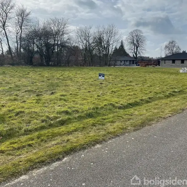 En grøn græsmark med et opsat til salg-skilt midt på marken. Baggrunden er en skov og en bygning. Tekst på skiltet: "Nybolig til salg."