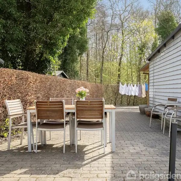 Et udendørs spisebord med træstole står på en flisebelagt terrasse. Et tørrestativ med hvide klæder ses i baggrunden. Omgivet af en hæk og et lyst træskur.