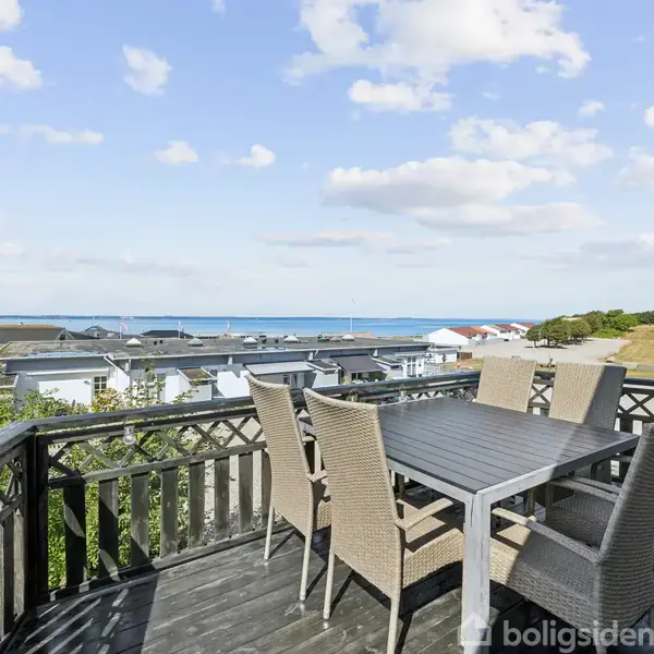Terrasse med havemøbler står stille, omgivet af en havudsigt, hustage og grønne marker under en blå, let skyet himmel.