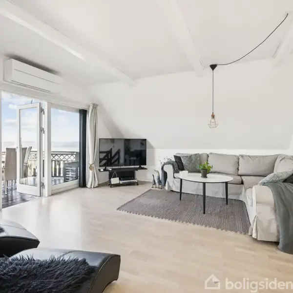 Stue med lys sofa og sofabord på tæppe. En fladskærms-TV står ved siden af åbne altandøre, der fører til en balkon med udsigt. Væggene og loftet er hvide.